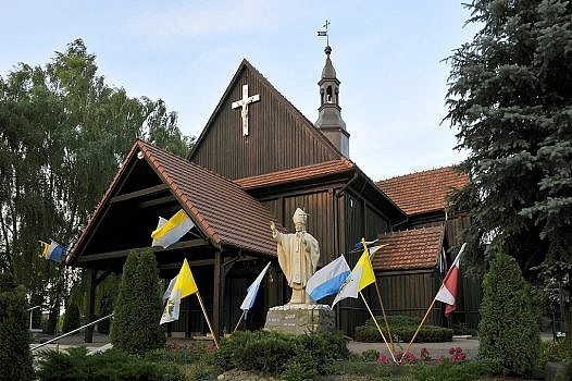 drewniany kościół w Chludowie widziany od frontu, na pierwszym planie pomnik Jana Pawła II w otoczenia flag biało-niebieskich i biało-żółtych