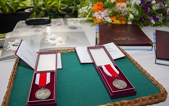 Dwa medale "Za zasługi dla obronności kraju" leżące na tacy, pod nimi legitymacje, za medalami teczki z listami gratulacyjnymi oraz kwiaty