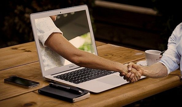 Laptop na stole, z ekranu którego ręka wychodzi na zewnątrz i wita się z osobą siedzącą przy stole