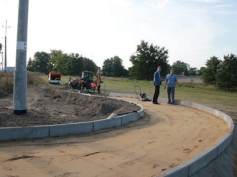 Widok na budowaną drogę po środku której stoją dwie osoby (na pierwszym planie), w tyle pracownicy i sprzęt drogowy.