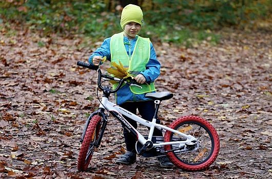 Dziecko stojące przy rowerze, ubrane w kamizelce odblaskowej, w jesiennej scenerii.