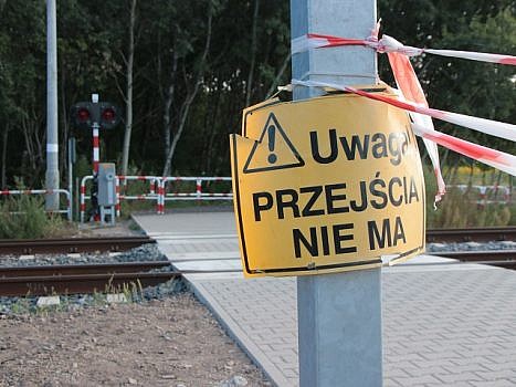 Słup, na którym przywieszona jest żółta tabliczka z ostrzeżeniem: uwaga przejścia nie ma. W tle tory kolejowe.