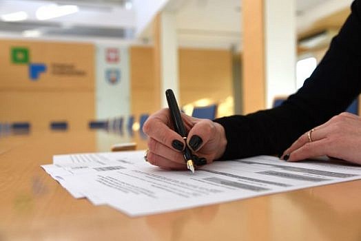 Zbliżenie na stół i ręka wypełniającą dokument. W tle logo powiatu poznańskiego.