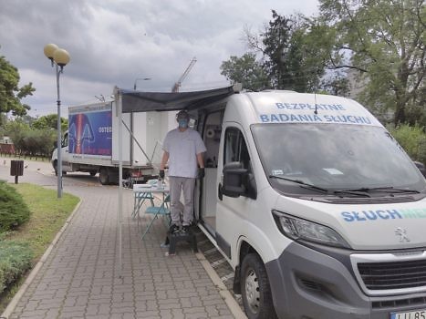 samochód typu van z napisem "bezpłatne badanie słuchu", przy aucie stoi mężczyzna w białym kitlu i maseczce chirurgicznej
