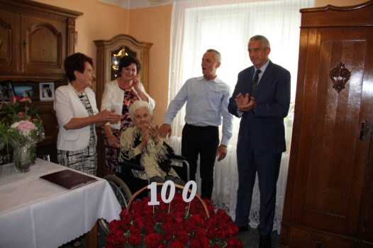 Jubilatka siedząca na wózku inwalidzkim przy stole w pokoju wraz z gośćmi w tym z wójtem Grzegorzem Wojterą