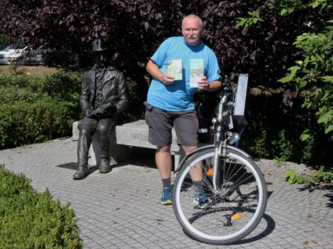 Pan Dariusz stojący przy rowerze na tle ławeczki z Wojciechem Bogusławskim w  Suchym Lesie. 