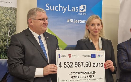 Wicemarszałek, stojący po prawej stronie zdjęcia, wspólnie z Prezes LGD, stojącą po lewej stronie zdjęcia, trzymają planszę z kwotą 4532987,60 euro, w tle roll up Suchego Lasu