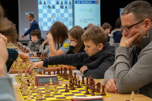 Szachiści, na pierszym planie stoły z szachownicami, w tle ekran z szachownicą online.