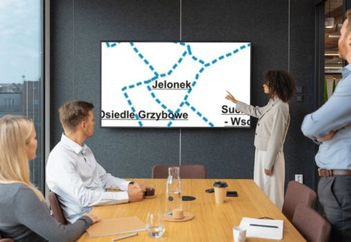 Zdjęcie sali ze stołem konferencyjnym, ludźmi siedzącymi i tablicą z mapą Jelonka oraz prowadzącą.