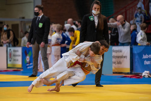 Dwóch zawodników judo walczy na macie, w tle dwóch sędziów i osoby przed trybunami.