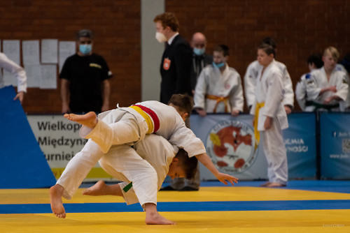 Walka judo, moment rzutu na matę, w tle inni zawodnicy i sędzie. 